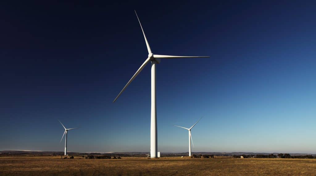 Three large wind turbines standing in an open field against a clear blue sky, with grassland visible in the foreground.