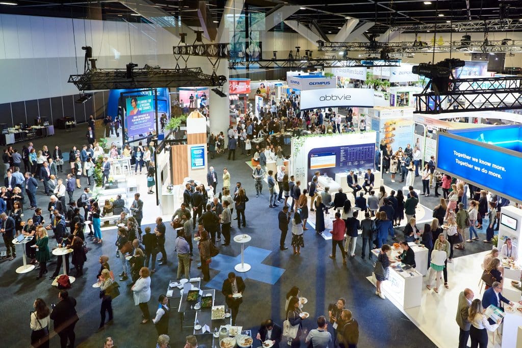 An aerial view of a large convention hall filled with people attending various booths and displays at an event. Brightly lit exhibits from different companies are set up around the space.
