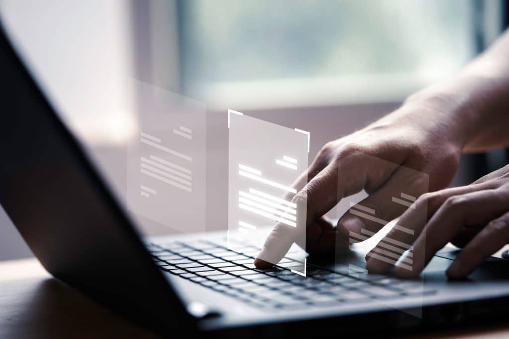 A person typing on a laptop keyboard with holographic documents displayed above the keyboard against a blurred background.