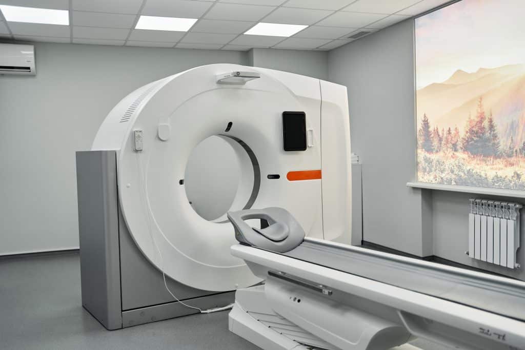 A modern CT scanner in a clean, bright medical room. The machine has a large circular opening and is positioned next to a patient bed. A window with a landscape view is visible in the background.