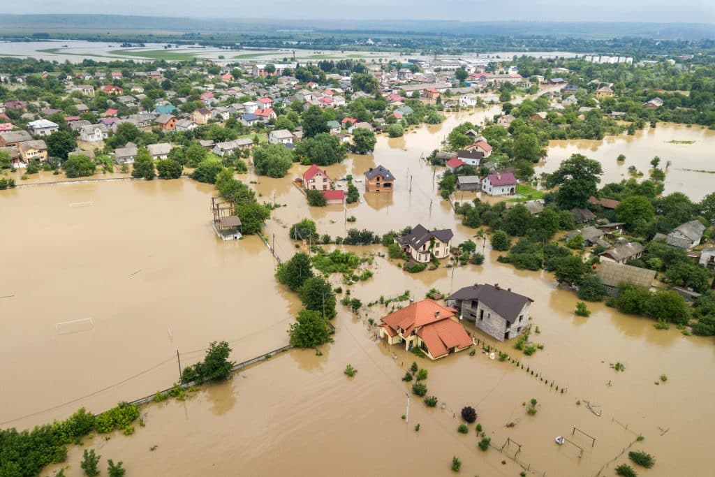 An aerial view of a flooded residential area, with houses partially submerged in water. Trees and roads are visible, while some structures remain above the flood level. Overcast sky and distant land can be seen in the background.
