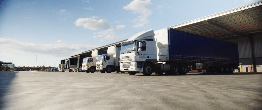 Several trucks parked in a loading area next to a warehouse. The trucks, including a large white semi-truck, are lined up on a paved surface, with a clear blue sky and clouds in the background.