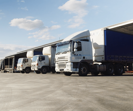 Several trucks parked in a loading area in front of a warehouse. The trucks are positioned in a row, with clear skies and scattered clouds visible in the background.