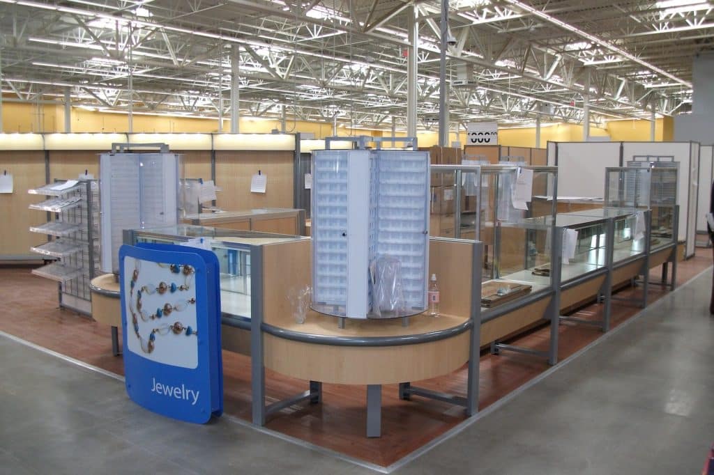 An empty jewelry display area inside a store featuring circular display cases, glass shelving units, and promotional signage indicating 'Jewelry'. The space has overhead lighting and a polished floor.