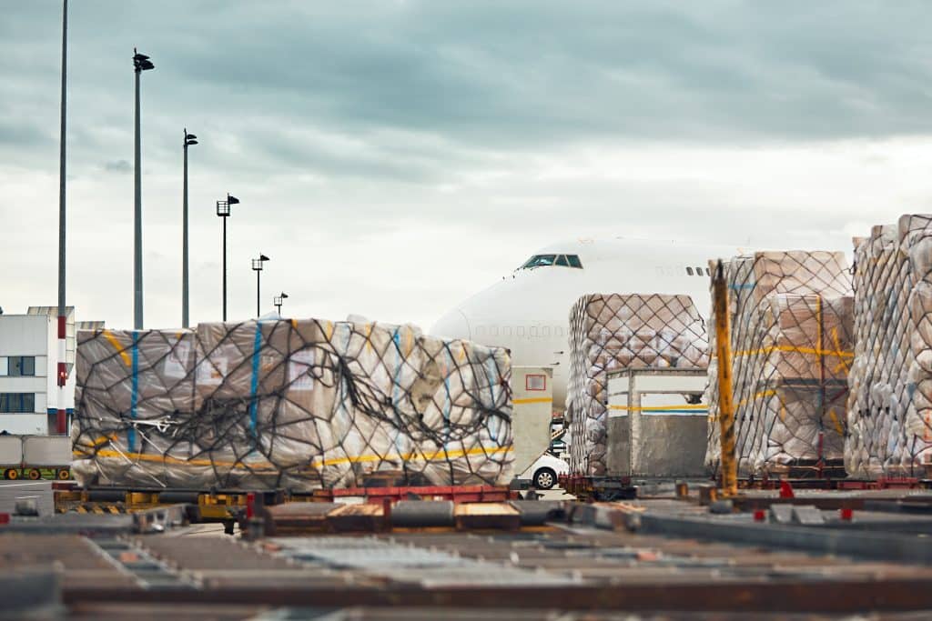 Cargo pallets stacked on a loading dock at an airport, with a large airplane visible in the background under a cloudy sky.