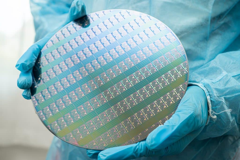 A person wearing blue gloves holds a shiny silicon wafer with multiple integrated circuit patterns, in a cleanroom environment.