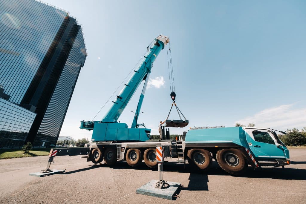 A blue crane lifting a heavy load at a construction site next to a modern glass building. Traffic cones are placed around the area for safety.