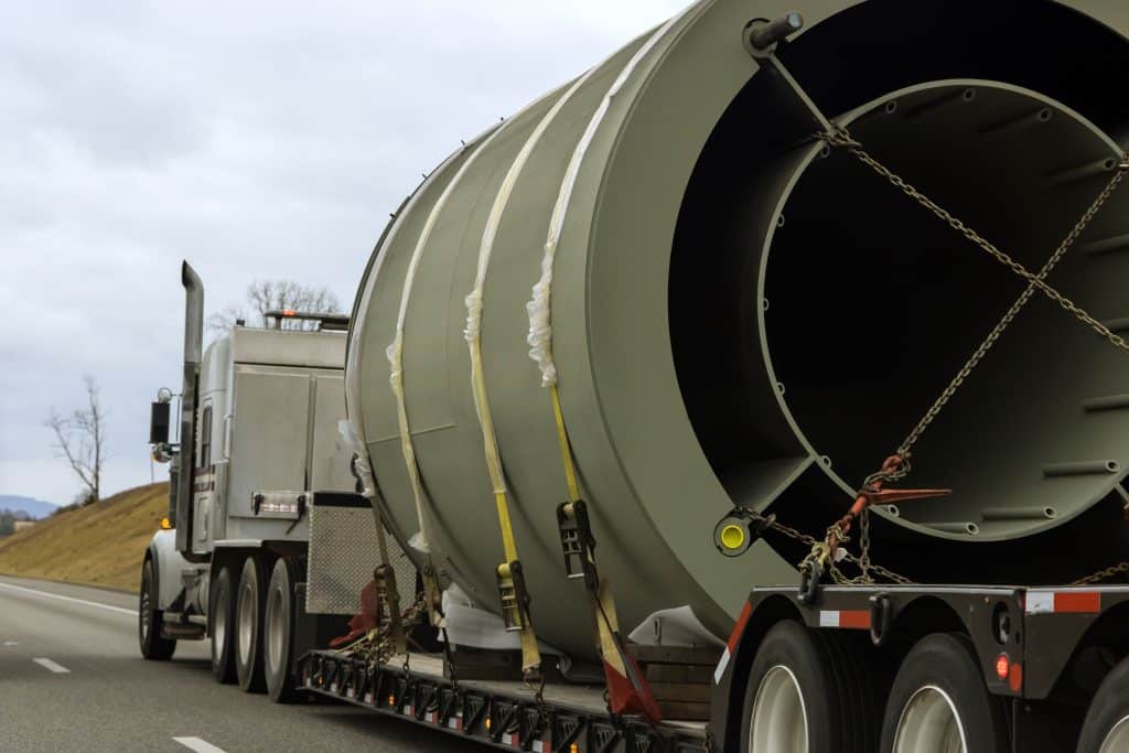 A large semi-truck transporting a cylindrical industrial object on a flatbed trailer. The truck is on a highway with a gray sky and trees in the background.