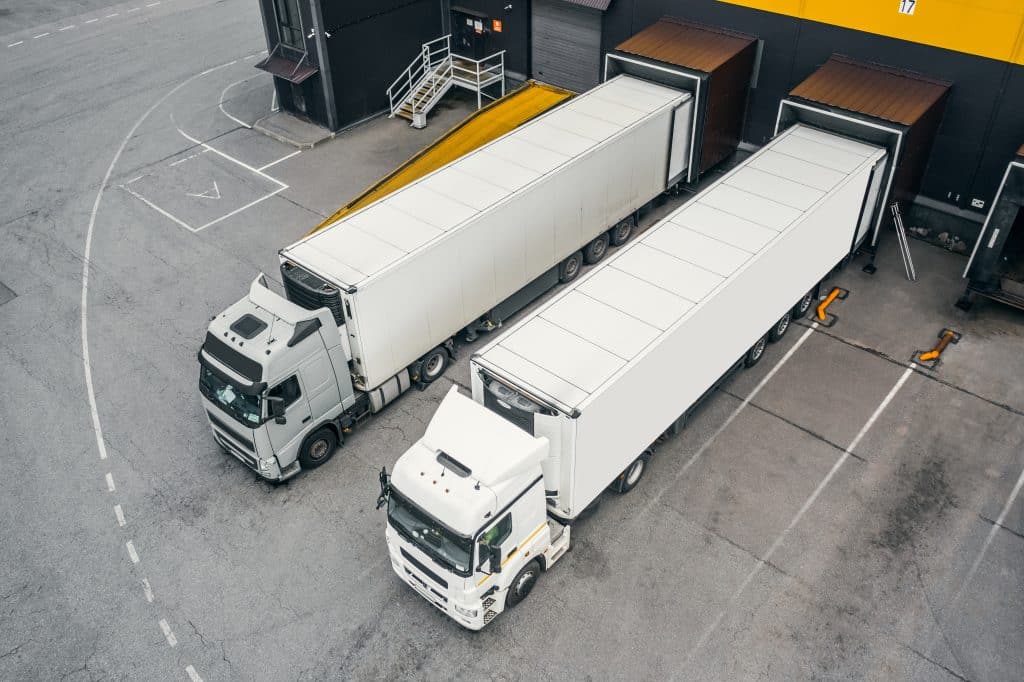 Two large trucks with white trailers parked in a loading dock area. One truck is facing the camera, while the other is positioned beside it. The loading dock has an industrial setting with a yellow overhang.