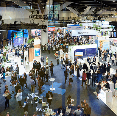An overhead view of a busy conference or exhibition hall filled with attendees interacting around various booths and displays, including digital screens and product presentations.